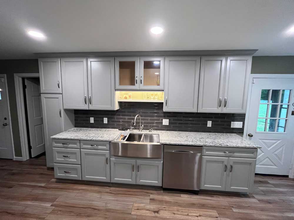 Kitchen sink area with grey cabinets, lighted shelf, granite counter tops and a tiled backsplash.