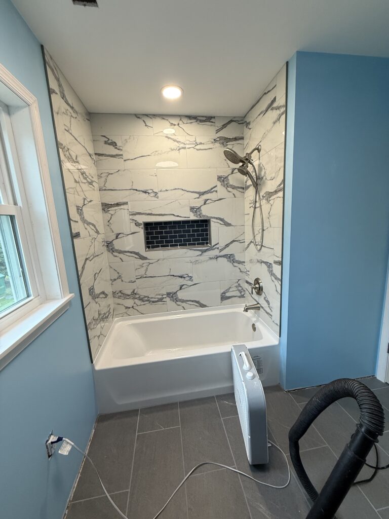 A modern bathroom with blue walls, a white bathtub, gray floor tiles, a marble-patterned shower wall, a recessed shelf with dark tiles, and a handheld showerhead.