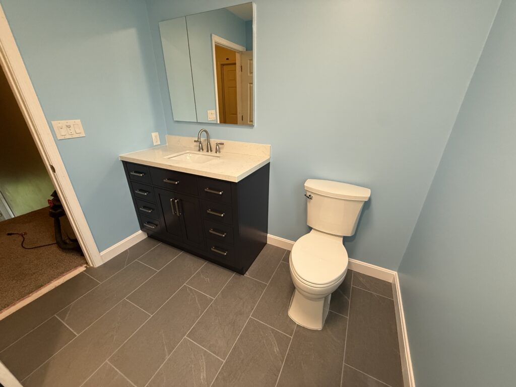 Dark blue bathroom vanity with a white quarts counter top and a white toilet. a glass recessed medicine cabinet. Light blue walls and a slate tiled floor.