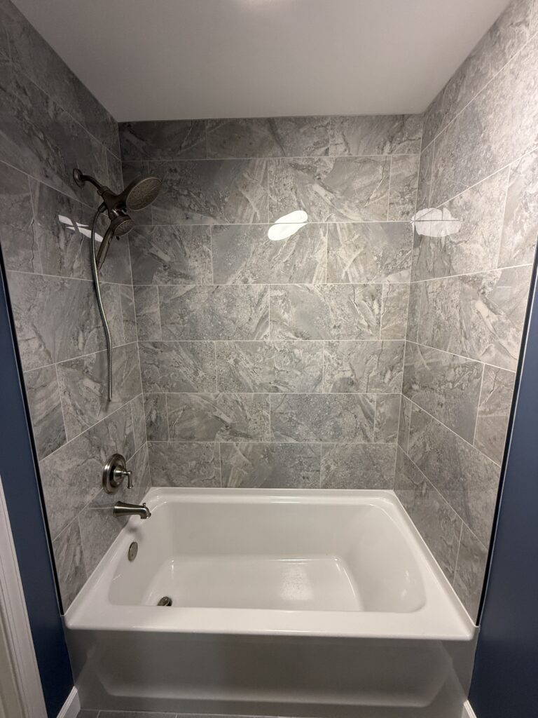 Modern bathroom with a white bathtub surrounded by gray marble-patterned tile walls. The shower features a brushed nickel showerhead with a handheld attachment and matching faucet fixtures. The surrounding wall outside the shower area is painted a deep blue.